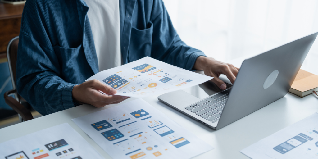 Person reviewing UX design mockups while working on a laptop, illustrating user experience design process in a modern workspace.